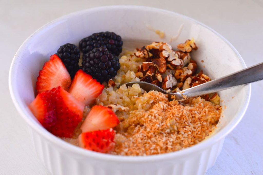 white bowl of quinoa topped with toasted coconut, chopped pecans, strawberries, and blackberries with spoon in it
