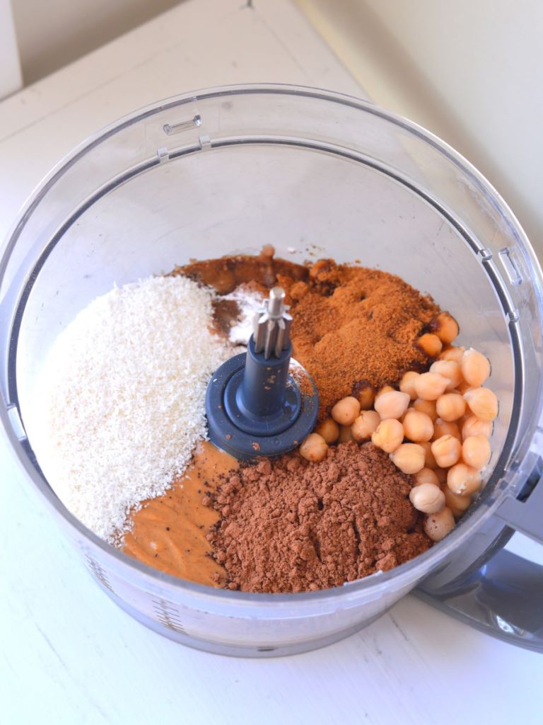 food processor bowl with unmixed chickpeas, coconut, coconut sugar, peanut butter, and other ingredients for cookies